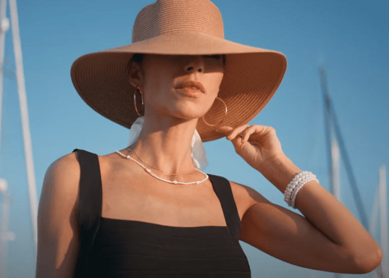 A woman wearing a wide-brimmed straw hat and black sleeveless dress stands outdoors. Her must-have accessories include hoop earrings, a necklace, and layered pearl bracelets, with the clear blue sky and sailboat masts in the background.